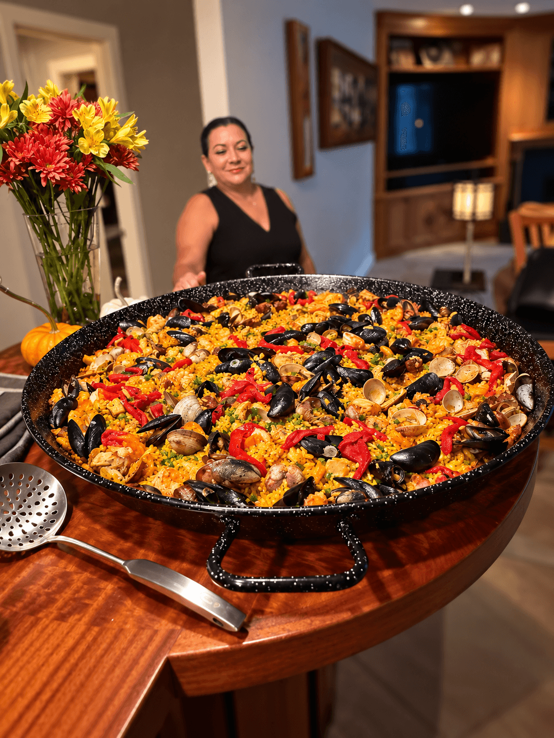 Close view of Gloria's Paella Mixta in a speckled pan, with Gloria visible softly in the background of her home.