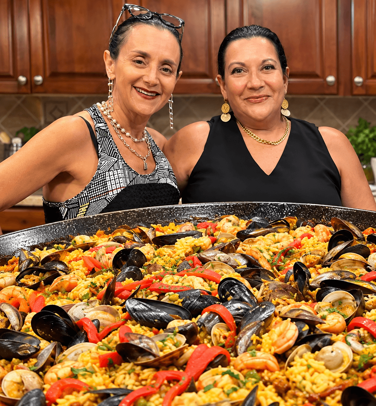 Gloria and her sister smiling together behind a large pan of freshly made Paella Mixta in their kitchen.